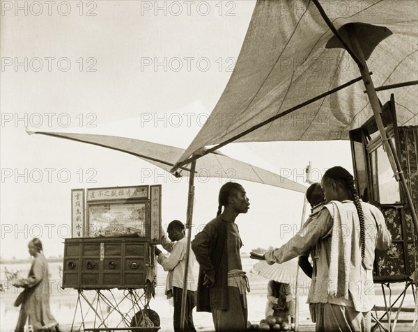 Peep boxes at a Chinese fair. Stallholders set up peep boxes beneath parasols at an outdoor fair. Each box was constructed with several small doors, that when opened, would reveal a unique miniature scene within. Probably China, circa 1910. China, People's Republic of, Eastern Asia, Asia.