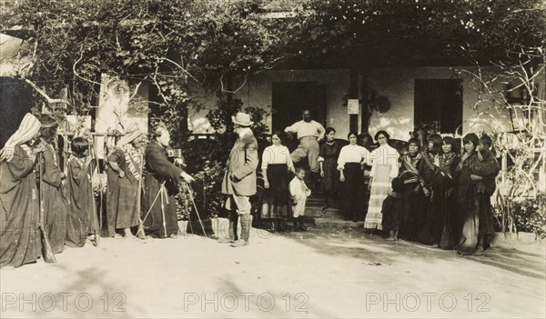 Photographing an indigenous group, Peru. Two European men set up a camera and tripod outside a colonial house, ready to photograph a group of indigenous Peruvian men, women and children wearing traditional dress. Several well-dressed members of the household watch the proceedings from the doorway. Peru, circa 1920. Peru, South America, South America .