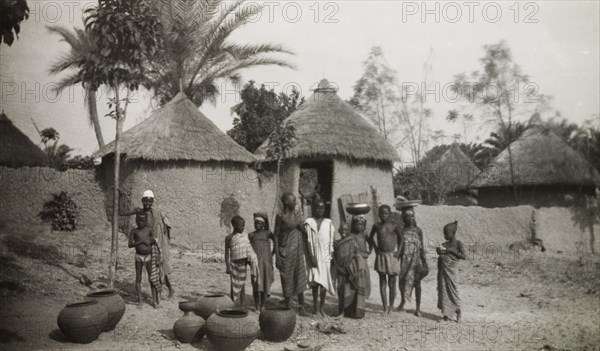 Young water carriers in Bida. A group of adolescents and children stand outside a cluster of mud-walled thatched huts beside clay pots used to collect water. An original caption comments: "..during the dry season (the village wells) dry up and the water has to be carried from the rivers and streams". Bida, Nigeria, circa 1955. Bida, Niger, Nigeria, Western Africa, Africa.