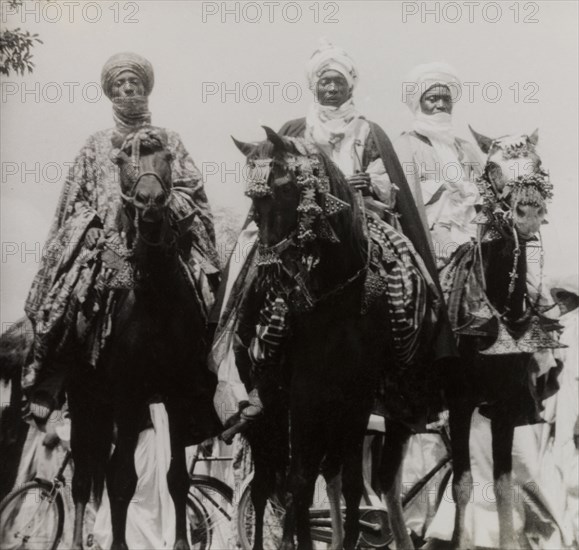 Nupe chiefs attend Sallah celebrations. Three Nupe chiefs dressed in ceremonial attire ride caparisoned horses during celebrations for Sallah, an Islamic event held to mark the end of Ramadan. Bida, Nigeria, October 1948. Bida, Niger, Nigeria, Western Africa, Africa.