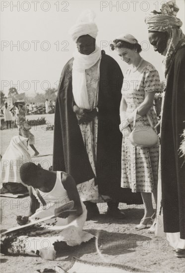 Queen Elizabeth II visits Kaduna Children's Village. Queen Elizabeth II is accompanied by the Sandauna of Sokoto and Makaman Bida (Regional Finance Minister) on a royal visit to Kaduna Children's Village. She smiles as she watches a young man demonstrate his writing skills outdoors. Kaduna, Nigeria, February 1956. Kaduna, Kaduna, Nigeria, Western Africa, Africa.