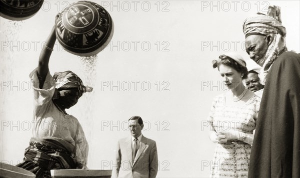 Queen Elizabeth II and Makaman Bida. Queen Elizabeth II and Makaman Bida (Regional Finance Minister) watch a demonstration of traditional domestic skills by young women at Kaduna Children's Village. The girls tip grain from bowls into clay pots, possibly wind-winnowing the chaff from wheat grain or rice. Kaduna, Nigeria, February 1956. Kaduna, Kaduna, Nigeria, Western Africa, Africa.