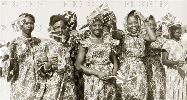 Flags for Queen Elizabeth II, Kaduna. Young women in traditional Nigerian dress wave union jack flags to welcome Queen Elizabeth II and the Duke of Edinburgh on their royal visit to Kaduna Children's Village. Kaduna, Nigeria, February 1956. Kaduna, Kaduna, Nigeria, Western Africa, Africa.