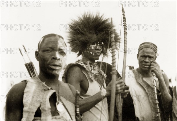 Nigerian hunters with bows and arrows. Portrait of three Nigerian hunters holding bows and arrows. They wear clothing made from animal skin and have their faces decorated with face paint. Nigeria, circa 1955. Nigeria, Western Africa, Africa.