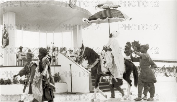 Durbar for Queen Elizabeth II, Nigeria. A Nigerian chief sits beneath a ceremonial umbrella as he rides a caparisoned horse past Queen Elizabeth II and the Duke of Edinburgh. The royal couple sit on thrones on a raised dais, attending a durbar held in honour of their visit. Kaduna, Nigeria, February 1956. Kaduna, Kaduna, Nigeria, Western Africa, Africa.