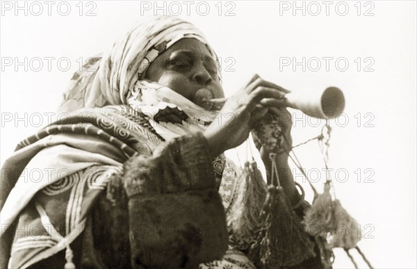 A Nigerian musician. A Nigerian musician plays a trumpet decorated with tassels. Nigeria, circa 1955. Nigeria, Western Africa, Africa.