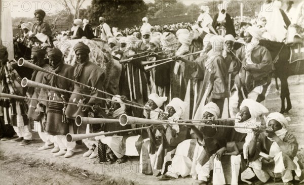 Nigerian trumpet players. A group of traditionally dressed Nigerian musicians assemble at an outdoor event to play a fanfare with long trumpets. Nigeria, circa 1955. Nigeria, Western Africa, Africa.