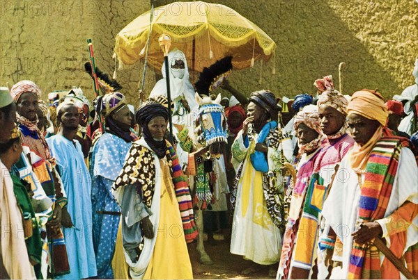 Alhaji Ado Bayero with his retinue. Portrait of Alhaji Ado Bayero (b.1930), the Emir of Kano, surrounded by members of his retinue who wear colourful ceremonial robes and head wraps. The Emir's face is covered and he sits beneath a ceremonial umbrella on the back of a caparisoned horse. Kano, Nigeria, circa 1975. Kano, Kano, Nigeria, Western Africa, Africa.