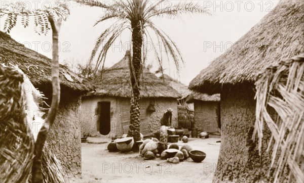 Interior of a Nigerian village. A woman undertakes domestic chores outdoors in the centre of a Nigerian village. The settlement comprises several round huts with mud walls and thatched roofs. Minna, Nigeria, circa 1940. Minna, Niger, Nigeria, Western Africa, Africa.