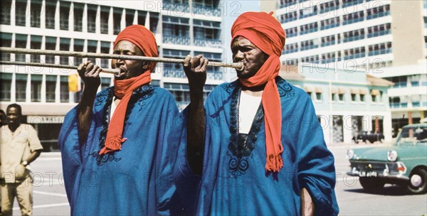 Trumpeters from North Nigeria'. Two musicians in traditional Nigerian dress play long trumpets on a modern city street. Northern Nigeria, circa 1965. Nigeria, Western Africa, Africa.