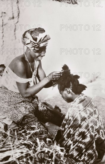 Hairdressing in Jos'. A Nigerian hairdresser smiles as she styles a customer's hair. Jos, Nigeria, circa 1940. Jos, Plateau (Nigeria), Nigeria, Western Africa, Africa.