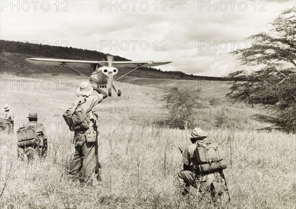 Kenya Police Airwing. Four officers of the Kenya Police Airwing watch as a light aircraft comes in to land on a grassy plain. Nakuru, Kenya, 1955. Nakuru, Rift Valley, Kenya, Eastern Africa, Africa.