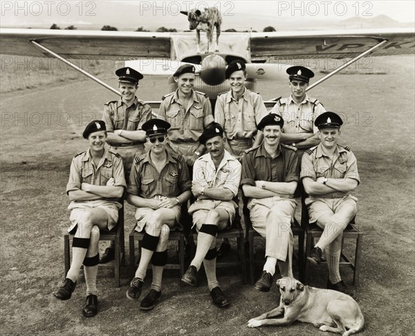 Kenya Police Airwing officers. Nine uniformed officers of the Kenya Police Airwing assemble for a group portrait with their dogs in front of a light aircraft at Mweiga operations base. Mweiga, Nyeri, Kenya, circa 1955. Nyeri, Central (Kenya), Kenya, Eastern Africa, Africa.
