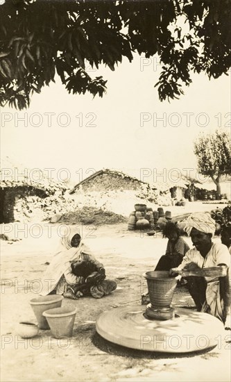 Indian craftsman at a potter's wheel. A craftsman sits beneath a shady tree as he sculpts a clay pot on a revolving wheel. India, circa 1920. India, Southern Asia, Asia.