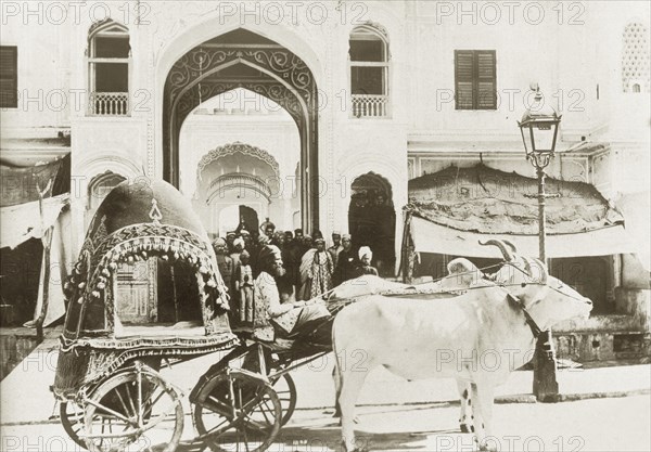 An ornate cattle-drawn carriage. An ornately covered carriage drawn by two bullocks waits outside a grand archway where a crowd of well-dressed Indian dignitaries are assembled. India, circa 1910. India, Southern Asia, Asia.