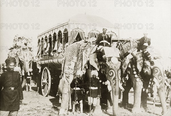 Procession of caparisoned elephants. Two caparisoned elephants pull a large, ornate carriage whilst a third carries a howdah. The animals have decorative patterns painted on their trunks and are ridden by 'mahouts' (elephant handlers). India, circa 1910. India, Southern Asia, Asia.