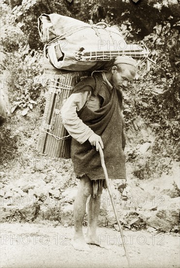 Burdened with luggage. An Indian man carries cumbersome bundles of luggage on his back as he walks barefoot along a road with the aid of a walking stick. India, circa 1920. India, Southern Asia, Asia.