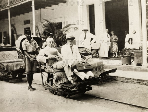 A rail trolley at Beira station. Two European men ride a narrow gauge rail trolley at Beira railway station, pushed by an African man in uniform. The trolley behind them displays the initials 'TZR', a reference the Trans-Zambeze Railway that ran between Beira and Nyasaland (Malawi). An original caption comments that this was "one method of transport before (the) car (and was) widely used". Beira, Mozambique, circa 1940. Beira, Sofala, Mozambique, Southern Africa, Africa.