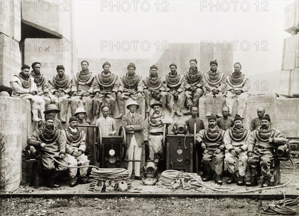 Construction engineers in diving suits. A team of British and Chinese civil engineers pose for a group portrait dressed in diving suits and surrounded by diving equipment. They were completing construction work at the Hong Kong docks under the supervision of Major Paul Dashwood (centre). Hong Kong, China, 1919. Hong Kong, Hong Kong, China, People's Republic of, Eastern Asia, Asia.