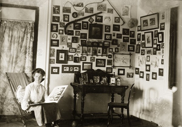 In the drawing room at Penshew House. A European woman browses through a photograph album in the drawing room of Penshew House. Behind her, the walls are covered with framed pictures and items of memorabilia. Bangalore, India, circa 1900. Bangalore, Karnataka, India, Southern Asia, Asia.