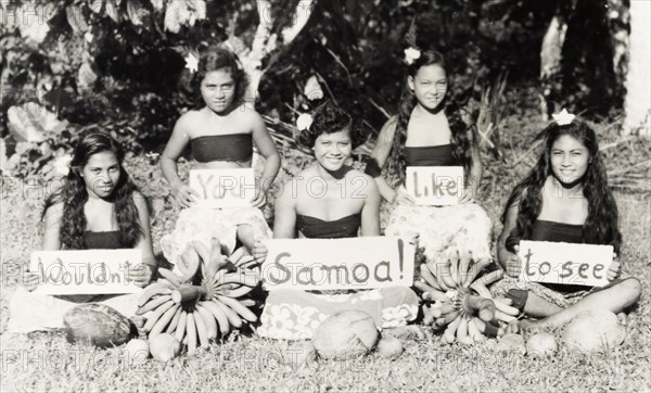 Wouldn't you like to see Samoa'. Five young Samoan women sit cross-legged on the grass, holding up signs that read 'Wouldn't you like to see Samoa'. The girls wear flowers in their hair and are surrounded by a display of locally produced fruit. Apia, Western Samoa (Samoa), circa 1956. Apia, Apia Urban Area, Samoa, Pacific Ocean, Oceania.