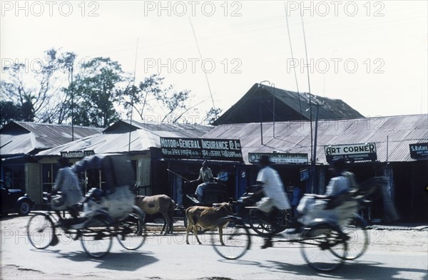 Cycle rickshaw drivers in Dibrugarh. Cycle rickshaw drivers whizz past shops with tin roofs on a commercial street in Dibrugarh. Dibrugarh, Assam, India, circa 1970. Dibrugarh, Assam, India, Southern Asia, Asia.