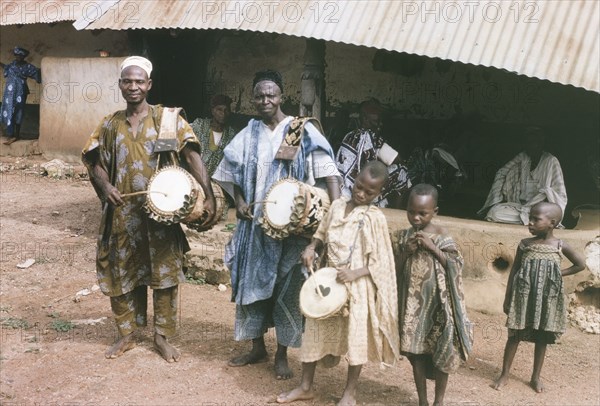 Musicians at Oyo. A group of adult and child musicians, originally captioned as 'palace drummers', perform on a city street. The two men play 'dunduns' (talking drums) whilst two of the children play a drum and a pipe. Oyo, Western Region (Oyo State), Nigeria, October 1963. Oyo, Oyo, Nigeria, Western Africa, Africa.