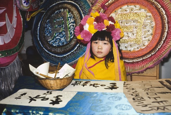 Chinese girl in a flower headdress. A young Chinese girl wears a headdress made of flowers as she sits at a script-writing stall during celebrations for the Chinese New Year. London, England, circa 1985. London, London, City of, England (United Kingdom), Western Europe, Europe .
