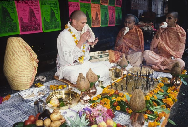 Hare Krishna stall at religious street festival. Members of the International Society for Krishna Consciousness, also known as the 'Hare Krishna' movement, sit at a stall crowded with flowers, fruit and vegetables, lit candles and incense sticks during a religious street festival. London, England, circa 1985. London, London, City of, England (United Kingdom), Western Europe, Europe .