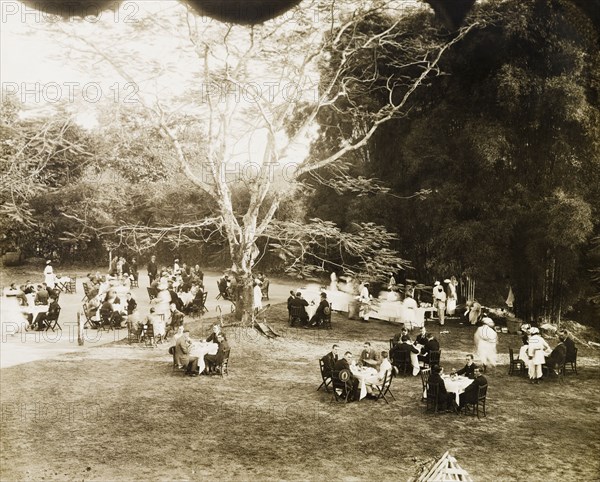A formal garden party. Small groups of European men and women sit around dining tables in the garden of a colonial residence, waiting to be served by Indian servants who dish out food from a long buffet table. India, circa 1925. India, Southern Asia, Asia.