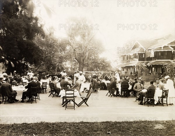 A formal garden party. Small groups of European men and women sit around dining tables on a tennis court in the grounds of a colonial residence, waiting to be served by Indian servants who dish out food from a long buffet table. India, circa 1925. India, Southern Asia, Asia.