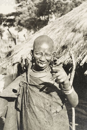 Portrait of a Kikuyu woman. Portrait of a smiling Kikuyu woman gesturing at the camera. Her head is shaven, indicating her status as a married woman, and she wears a number of hooped earrings. Kenya, circa 1934. Kenya, Eastern Africa, Africa.