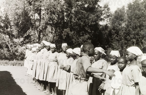 Kikuyu students at a mission school. Young Kikuyu women are lined up outdoors at the Church of Scotland mission school. An original caption comments that "the girls are dressed in European clothing and taught domestic skills". South Nyeri, Kenya, 1936. Nyeri, Central (Kenya), Kenya, Eastern Africa, Africa.