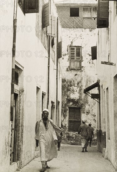 A back street of Lamu. A man in traditional Arab dress, including a kurta (shirt) and a fez, walks along a narrow residential back street in Lamu. Lamu, Kenya, circa 1947. Lamu, Coast, Kenya, Eastern Africa, Africa.