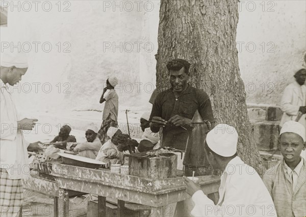 Snack food in Lamu. A street trader grills snack food in Lamu's main square. Lamu, Kenya, circa 1947. Lamu, Coast, Kenya, Eastern Africa, Africa.