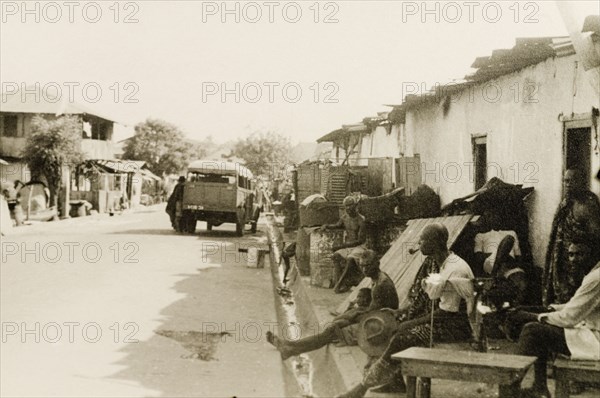 A side street in Accra. A concrete drainage ditch runs along a side street in Accra, where a group of men lounge on furniture cluttering the pavement outside a row of single storey buildings. Accra, Gold Coast (Ghana), circa 1950. Accra, East (Ghana), Ghana, Western Africa, Africa.