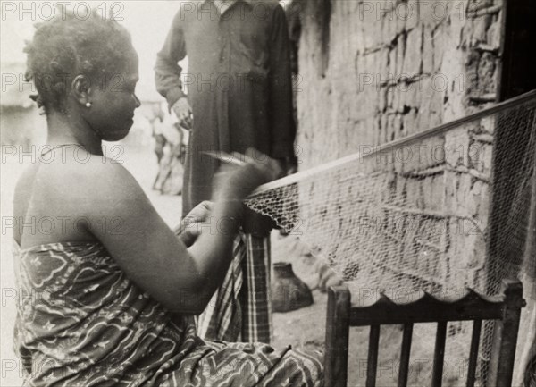 Repairing fishing nets. A Ghanaian woman in traditional dress sits outside a mud-walled building as she repairs fishing nets. Gold Coast (Ghana), circa 1950. Ghana, Western Africa, Africa.