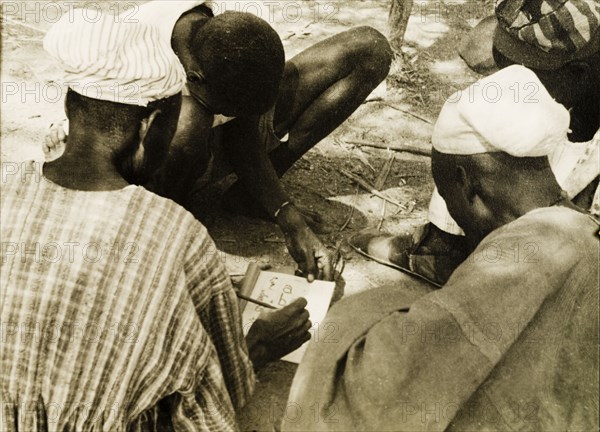 Adult literacy campaign, Gold Coast. A student teacher helps men to read from a book during a literacy class: part of a mass literacy campaign launched by the British government in 1951 to improve literacy skills amongst African adults, both in their own languages and in English. Northern Territories, Gold Coast (Northern Ghana), circa 1951., North (Ghana), Ghana, Western Africa, Africa.