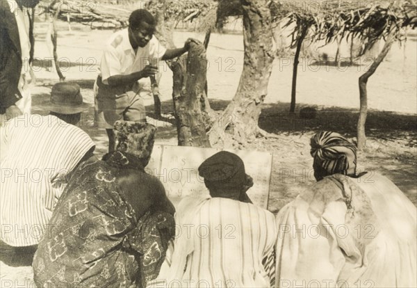 Adult literacy campaign, Gold Coast. A student teacher helps men to read from a board during a literacy class: part of a mass literacy campaign launched by the British government in 1951 to improve literacy skills amongst African adults, both in their own languages and in English. Northern Territories, Gold Coast (Northern Ghana), circa 1951., North (Ghana), Ghana, Western Africa, Africa.
