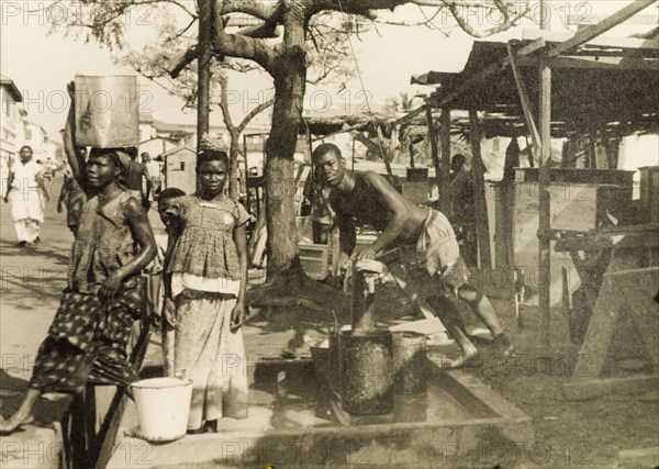 Collecting water from a hand pump, Accra. Men and women collect water from a hand pump on a street in Accra. Accra, Gold Coast (Ghana), circa 1950. Accra, East (Ghana), Ghana, Western Africa, Africa.