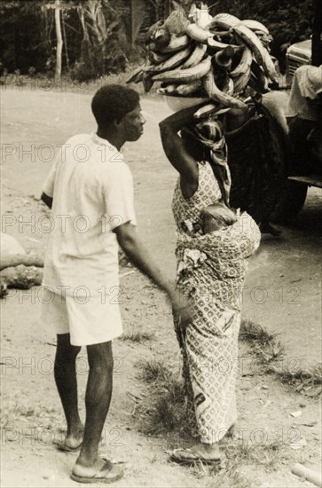 Gold Coast courtesy'. A man helps his wife to arrange her load as she attempts to balance a bowl of plantains on her head. Gold Coast (Ghana), circa 1950. Ghana, Western Africa, Africa.