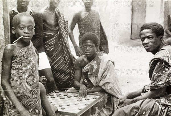 Asante boys playing a board game. A group of Asante (Ashanti) youths gather round to watch two boys play a board game on a street corner. Asante, Gold Coast (Ghana), circa 1950., Ashanti, Ghana, Western Africa, Africa.