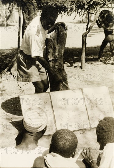 Adult literacy campaign, Gold Coast. A student teacher helps men to read from a board during a literacy class: part of a mass literacy campaign launched by the British government in 1951 to improve literacy skills amongst African adults, both in their own languages and in English. Northern Territories, Gold Coast (Northern Ghana), circa 1951., North (Ghana), Ghana, Western Africa, Africa.