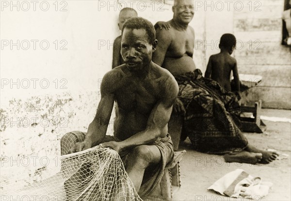 Fisherman repairing his nets, Ghana. A fisherman sits on a stool at the side of a street as he repairs his nets. Accra, Gold Coast (Ghana), circa 1950. Accra, East (Ghana), Ghana, Western Africa, Africa.