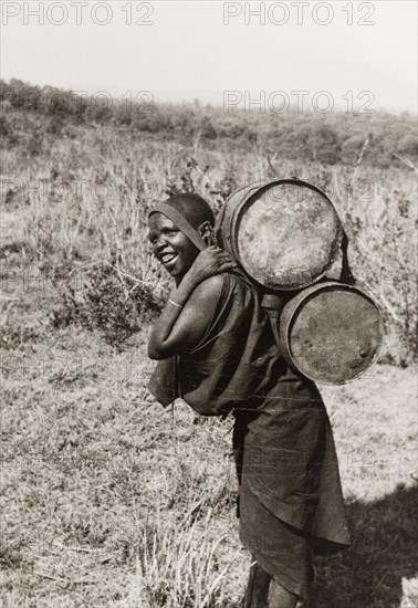 Kikuyu woman carrying water. A Kikuyu woman carries water in two barrels on her back, supported by a strap across her forehead. Kenya, circa 1932. Kenya, Eastern Africa, Africa.