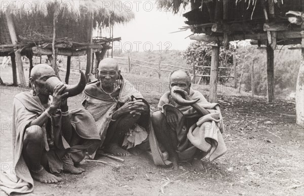 Kikuyu elders drinking beer. Three Kikuyu elders squat on the ground as they drink beer from hollow bullock horns, a privilege granted only to tribal elders. South Nyeri, Kenya, 1936. Nyeri, Central (Kenya), Kenya, Eastern Africa, Africa.