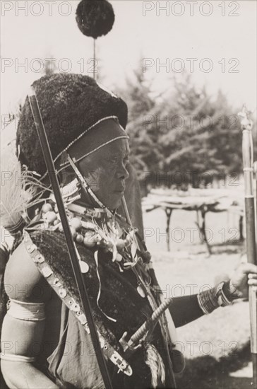 Kikuyu chief in ceremonial dress. Profile shot of a Kikuyu chief wearing full ceremonial dress. He wears strings of necklaces made from bone and beads and a fur headdress over a cap made from a sheep's stomach. South Nyeri, Kenya, 1936. Nyeri, Central (Kenya), Kenya, Eastern Africa, Africa.
