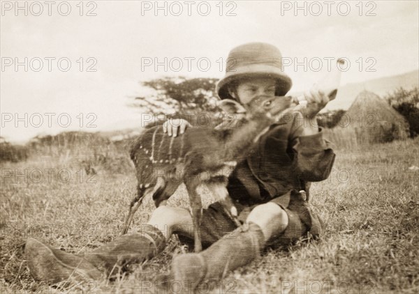 Elspeth Grant feeds an antelope calf. Elspeth Grant (later Huxley) sits on the grass feeding milk to her pet antelope calf Twinkle. Her settler farmer parents, Jos and Nellie Grant, had arrived in Kenya in 1912 to set up a coffee farm at Thika. Elspeth joined them at the family's new home, Kitimura Estate, on New Year's Day 1914. Thika, Kenya, circa 1914. Thika, Nairobi Area, Kenya, Eastern Africa, Africa.