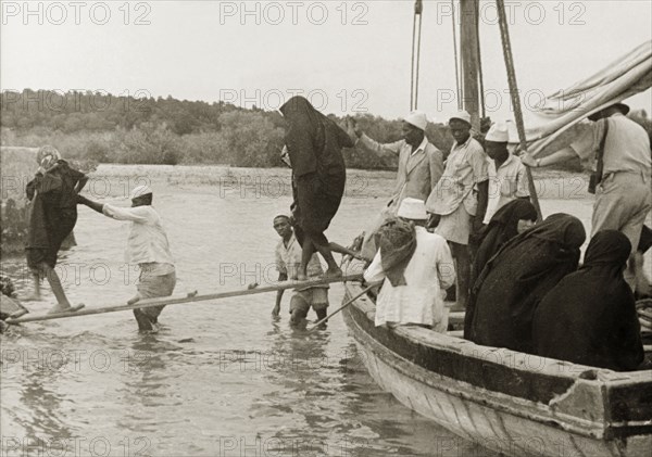 Disembarking at Mombasa Island. Arabic men assist a group of veiled Muslim women to cross a gangplank as they disembark from a sailing boat at Mombasa Island. Mombasa, Kenya, circa 1935. Mombasa, Coast, Kenya, Eastern Africa, Africa.