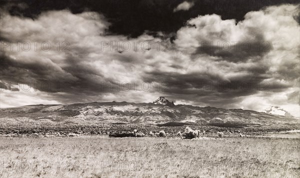 Ominous clouds over Mount Kenya. View across a grassy landscape looking towards Mount Kenya, where ominous clouds hang over Batian Peak. Central Kenya, circa 1935., Central (Kenya), Kenya, Eastern Africa, Africa.